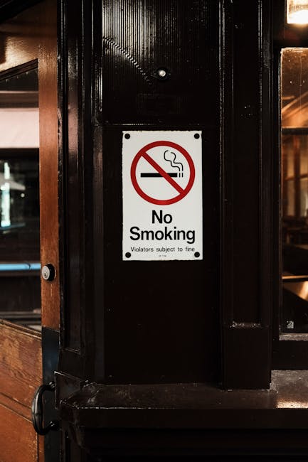 Close-up image of a rectangular 'No Smoking' sign affixed to a black-painted wooden or metal pillar inside an indoor setting, possibly a restaurant or café. The sign features a red circle and diagonal line crossing out a stylized cigarette icon, with the words 'No Smoking' printed in black underneath. Small text below reads 'Violators subject to fine.' The background shows blurred interior elements, including part of a wooden or laminate wall and a glass window or door with reflections, suggesting a warm ambient light and a cozy atmosphere. The sign's placement indicates an effort to enforce a non-smoking policy in an enclosed space, reflecting typical interior signage related to public health and safety regulations concerning waste management and cleanliness. The overall scene emphasizes the importance of clear communication about restricted behaviors that can contribute to indoor environmental quality, relevant to practices of responsible waste and health regulation compliance.