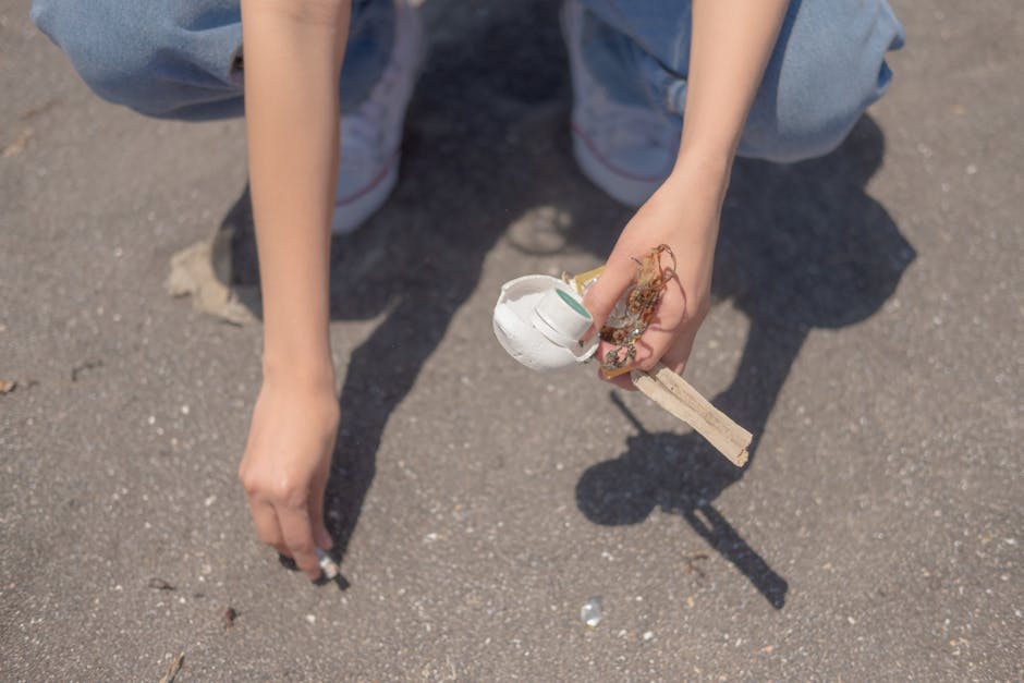 A close-up view of a person's hands and arms, with the individual crouching on a paved surface, possibly a driveway or sidewalk. One hand holds a small, white, partially broken plastic container or cup, with some debris and tangled fibers attached to it. The person's clothing includes light blue jeans and white sneakers with faint red and blue accents. The surface beneath is coarse, gray asphalt with small scattered stones and faint shadow cast by the person's arms and the object. The background is indistinct, focusing solely on the hands and the object being held, highlighting an act of littering or debris collection that relates to waste management and rubbish removal services typically observed in private or professional clearance scenarios.