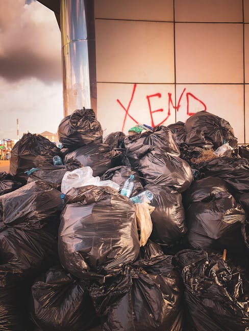 A large pile of black plastic rubbish bags is stacked against a tiled wall with beige and peach tones, situated outdoors on what appears to be a pavement or concrete surface. The bags are bulging and tied at the top, containing discarded waste, with some partially torn open revealing plastic bottles and other debris. Behind the bags, graffiti with the word 'XEND' in red spray paint is visible on the wall. The scene is lit by natural light, casting soft shadows on the bags and the surrounding ground, emphasizing the disorder of the refuse. The environment suggests an area where private waste disposal or on-site rubbish clearance might be necessary, and the presence of the graffiti hints at potential for independent rubbish removal services, such as those offered by Rubbish Clearance Haringey. This image visually underscores the need for effective rubbish removal solutions in urban settings, especially in contexts where regular council bin collection may not be available or sufficient.