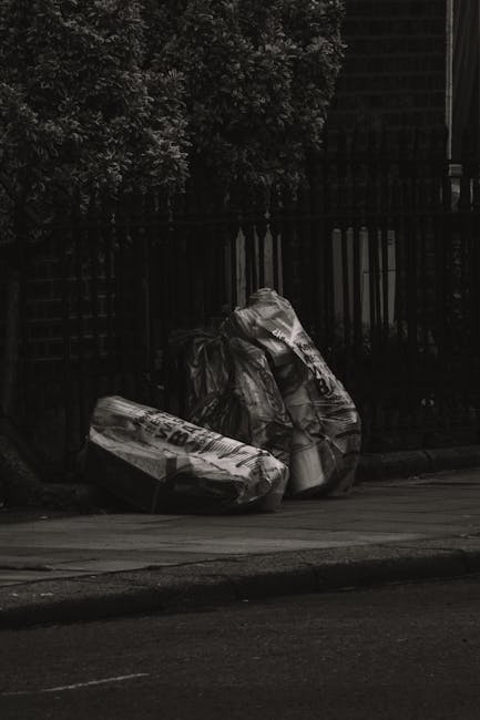 A close-up view of a person's hands and arms, with the individual crouching on a paved surface, possibly a driveway or sidewalk. One hand holds a small, white, partially broken plastic container or cup, with some debris and tangled fibers attached to it. The person's clothing includes light blue jeans and white sneakers with faint red and blue accents. The surface beneath is coarse, gray asphalt with small scattered stones and faint shadow cast by the person's arms and the object. The background is indistinct, focusing solely on the hands and the object being held, highlighting an act of littering or debris collection that relates to waste management and rubbish removal services typically observed in private or professional clearance scenarios.