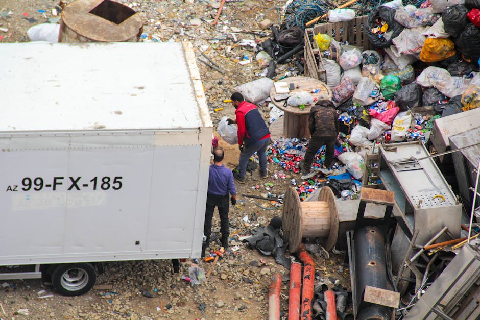 A high-angle view of a large outdoor area cluttered with assorted waste and rubbish, including plastic bags, cardboard boxes, and discarded household items. In the center, three individuals are actively engaged in debris collection, with one person wearing a red top and dark trousers, bending over to pick up items. Around them, there are various damaged furniture pieces, spools, and construction materials such as pipes and metal fragments. To the left, part of a white truck with black alphabetic and numeric markings is visible, which appears to be used for rubbish disposal. The ground is uneven, covered with dirt, small rocks, and scattered trash, conveying a scene of on-site removal or clearing of accumulated waste, likely handled by an independent waste disposal service like Rubbish Clearance Haringey. The environment suggests a non-domestic, possibly industrial or vacant lot setting, where rubbish collection is actively taking place amidst the cluttered surroundings.