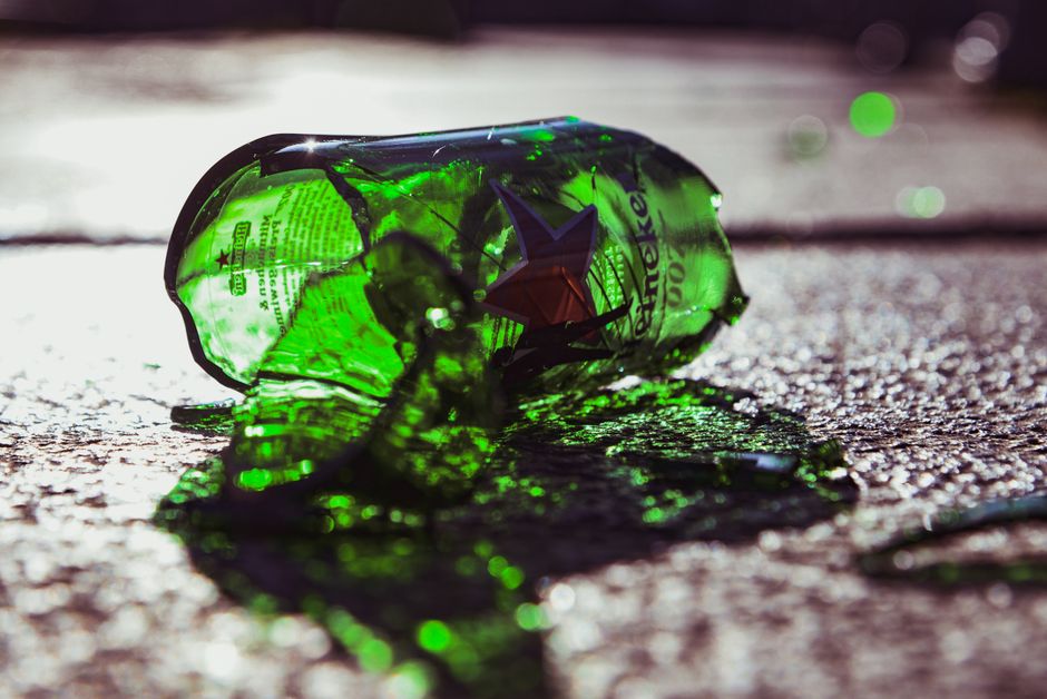 A crushed green glass bottle lies on a paved surface outdoors, casting a shadow in bright natural light. The bottle's label is partially visible with printed text, and its surface exhibits cracks and sharp edges typical of broken glass. The surrounding area includes the textured pavement with small pebbles and dirt, and the background is blurred with additional reflections and indistinct shapes. The scene captures an incident of discarded waste that could be addressed through private rubbish clearance services, emphasizing the importance of proper waste disposal. The natural sunlight highlights the translucent quality of the broken glass, and the overall composition reflects an environment where waste is improperly disposed of, potentially requiring intervention such as rubbish removal from public or private properties. Rubbish Clearance Haringey’s expertise in handling such debris aligns with the scene, illustrating the need for efficient rubbish collection and clear-up services to maintain cleanliness and safety in shared outdoor spaces.