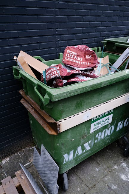A green wheelie bin filled with red and black plastic bags containing building materials, placed on a paved surface against a dark brick wall. The bags are labeled with specifications and brand names, indicating they contain construction or renovation debris. The bin is situated outdoors next to another similar bin, and a metal ramp or grate is leaning against the side of the bin. The scene depicts a typical private waste disposal setup, possibly related to site clearance or DIY project waste, with the presence of branded bags and the bin's positioning suggesting on-site clearance rather than public waste collection, aligning with alternative rubbish removal options that might be addressed by services like Rubbish Clearance Haringey.