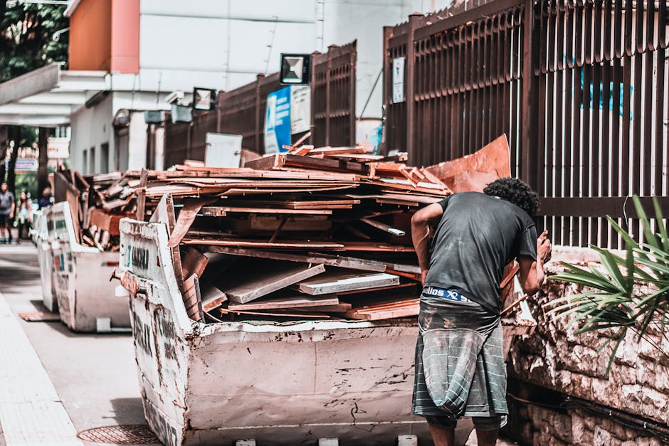 A man with curly black hair, wearing a dark grey T-shirt and plaid shorts, is leaning over a large, white, industrial skip filled with various construction debris, including broken wooden panels, tiles, and other building materials. The skip is positioned on the pavement beside a brown metal fence with vertical slats, which runs along the edge of the sidewalk. In the background, there are signs, a building with a white exterior and a small orange section, and a few pedestrians walking further along the street. The scene is set outdoors, illuminated by natural daylight, highlighting the textures of the debris and the weathered surface of the skip. This image illustrates a typical scenario of on-site rubbish clearance, possibly related to private waste disposal or building renovation waste removal, managed by companies such as Rubbish Clearance Haringey, emphasizing the importance of proper rubbish handling outside conventional council collection services.