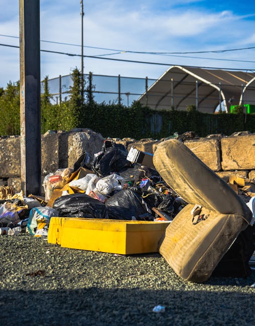 A close-up view of a curbside collection of mixed household waste positioned against a weathered stone wall in an outdoor area, with discarded black and transparent rubbish bags containing various refuse, including plastic packaging and paper. In the foreground, an old, stained, light-colored upholstered armchair with visible wear and tear is tilted on its side, partially overlapping the waste. A yellow plastic tray and an unidentified large, curved piece of debris are also situated among the waste. Behind the heap, a metal pole supports overhead cables, and in the background, a chain-link fence separates the area from a green hedge, with a large tent-like structure with curved roof panels and a partially visible electronic display to the right. The scene is lit by natural daylight, creating shadows and highlights on the rubbish, and conveys a typical example of private rubbish disposal near a residential or commercial property, illustrating the importance of proper waste management and collection service options, such as those offered by Rubbish Clearance Haringey.