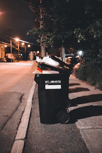 A black wheeled rubbish bin positioned on the pavement next to a curb on a dimly lit residential street at night. The bin is labeled with the name 'St. John's' and filled with assorted waste, including crumpled paper, cardboard boxes, and plastic bags, some of which are visible over the open lid. The surrounding environment features a row of trees and shrubbery along the sidewalk, with streetlights casting warm illumination on the scene. In the background, there are parked cars and distant building lights. The scene suggests a private or independent waste collection process, typical of local rubbish removal services like those offered by Rubbish Clearance Haringey, which handle waste outside of municipal collection schedules. The overall atmosphere emphasizes waste management in residential areas, highlighting the importance of proper rubbish disposal and collection solutions for domestic and commercial properties.
