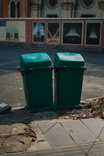 Two green plastic rubbish bins with hinged lids are placed side by side on a concrete pavement at the edge of a street. The bins are slightly weathered, with visible scratches and some dirt. They are positioned on a small patch of ground with scattered dry leaves and debris around their base. Behind the bins, there is an asphalt road and a decorative brick wall with painted advertisements or posters, partially visible at the top of the image. The scene appears to be in an urban area, with sunlight casting slight shadows, indicating a clear day. The image suggests an example of private waste collection or on-site rubbish disposal, which may be handled independently by local residents or waste management services such as Rubbish Clearance Haringey. The overall setting emphasizes the importance of proper rubbish storage and disposal in urban environments for effective waste management and recycling efforts.