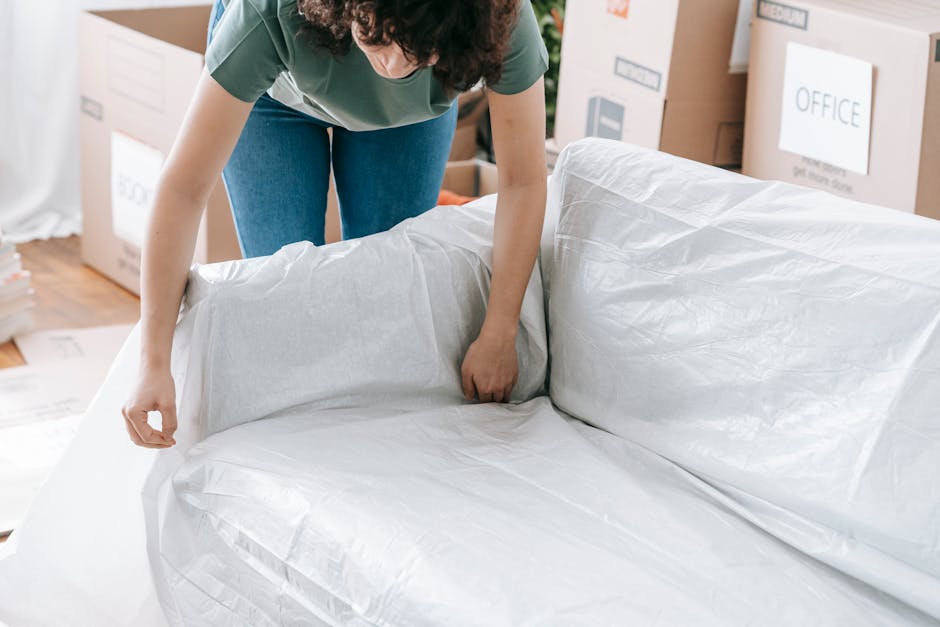 A person with curly dark hair, dressed in a green top and blue jeans, is in the process of wrapping a large, white, plastic-covered mattress or cushion with protective material. The individual is bending over, using both hands to secure the covering, which appears to be a moving or disposal wrap. The scene is set indoors, likely in a home or storage area, with multiple cardboard boxes visible in the background, some labeled with household or office-related tags such as 'OFFICE' and 'MEDIUM'. The boxes are stacked or placed against the walls, indicating an ongoing room clearance, possibly ahead of a house move or renovation. The wooden floor beneath is partially visible, and natural light illuminates the space, creating a neutral, functional atmosphere suitable for efficient rubbish or waste removal activities. Rubbish Clearance Haringey specializes in private waste collection, including large household items, and the scene depicts part of an on-site clearance process involving wrapping and preparing items for disposal or recycling, aligning with tailored waste management services.